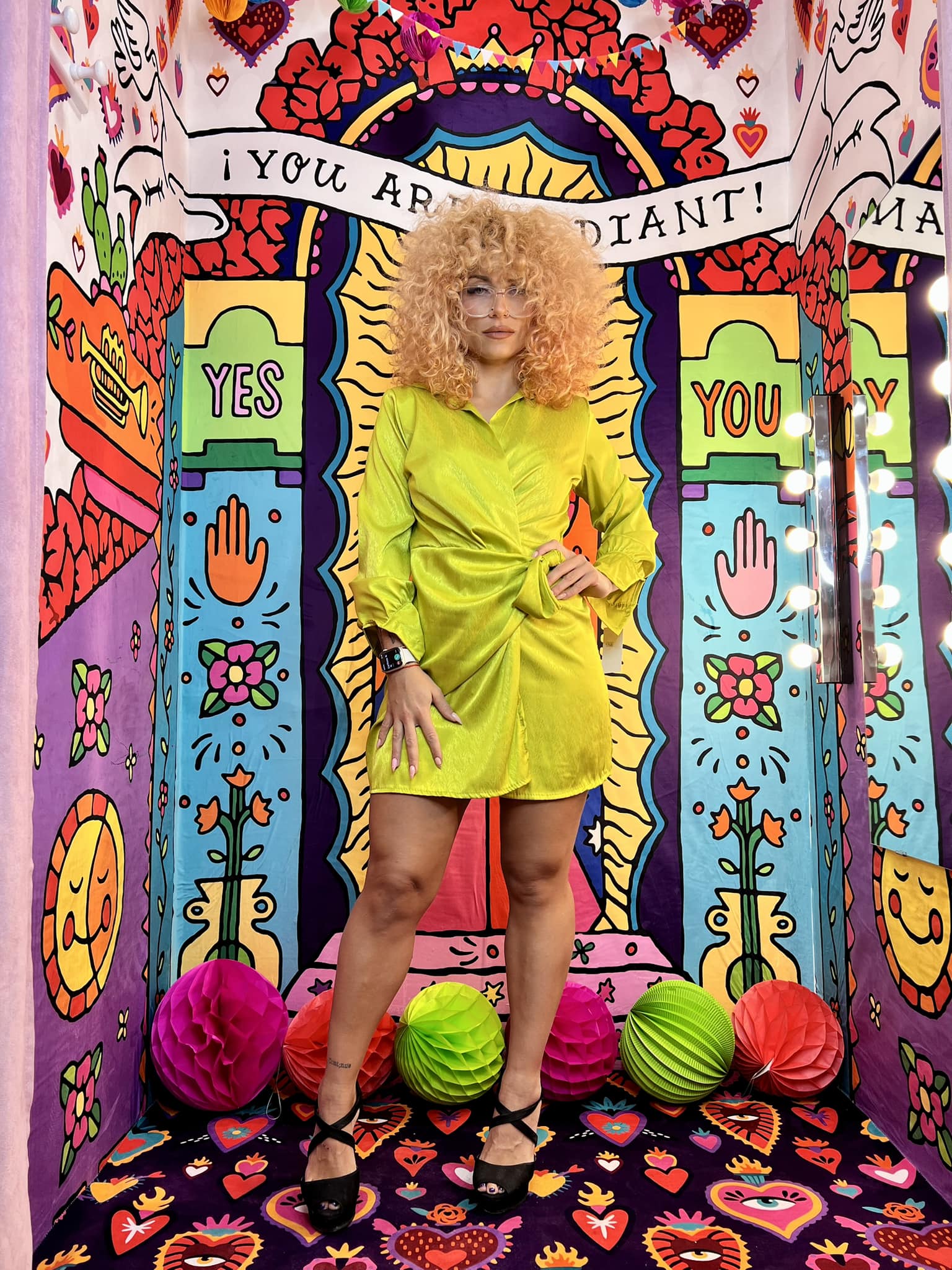 Foto de la nueva campaña publicitaria de la tienda Modelo rubia con vestido corto amarillo de satén posando frente a un mural colorido